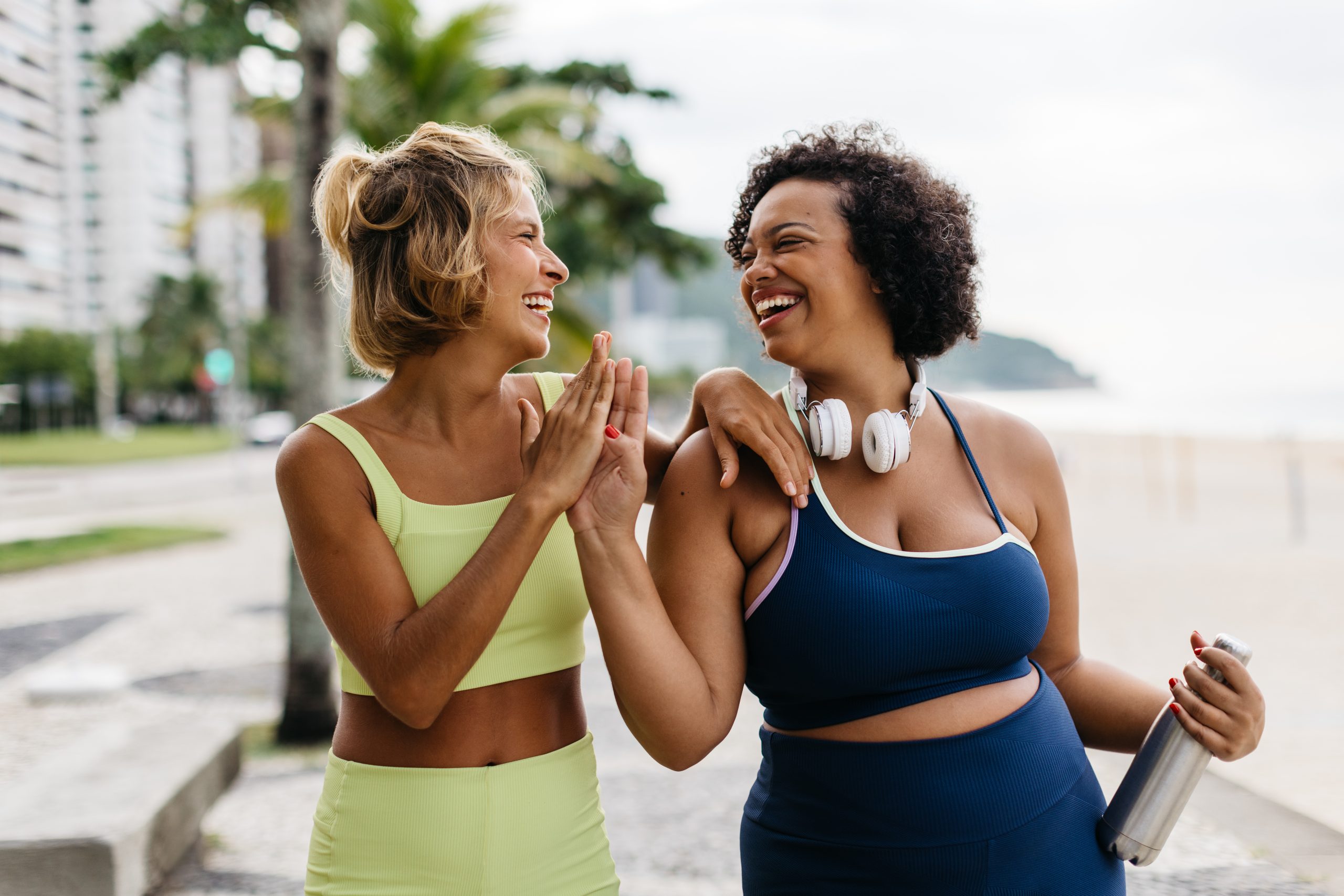 Sporty women high fiving each other in fitness wear, celebrating a successful workout session outdoors. Two fitness friends with different body sizes embrace a healthy lifestyle together.